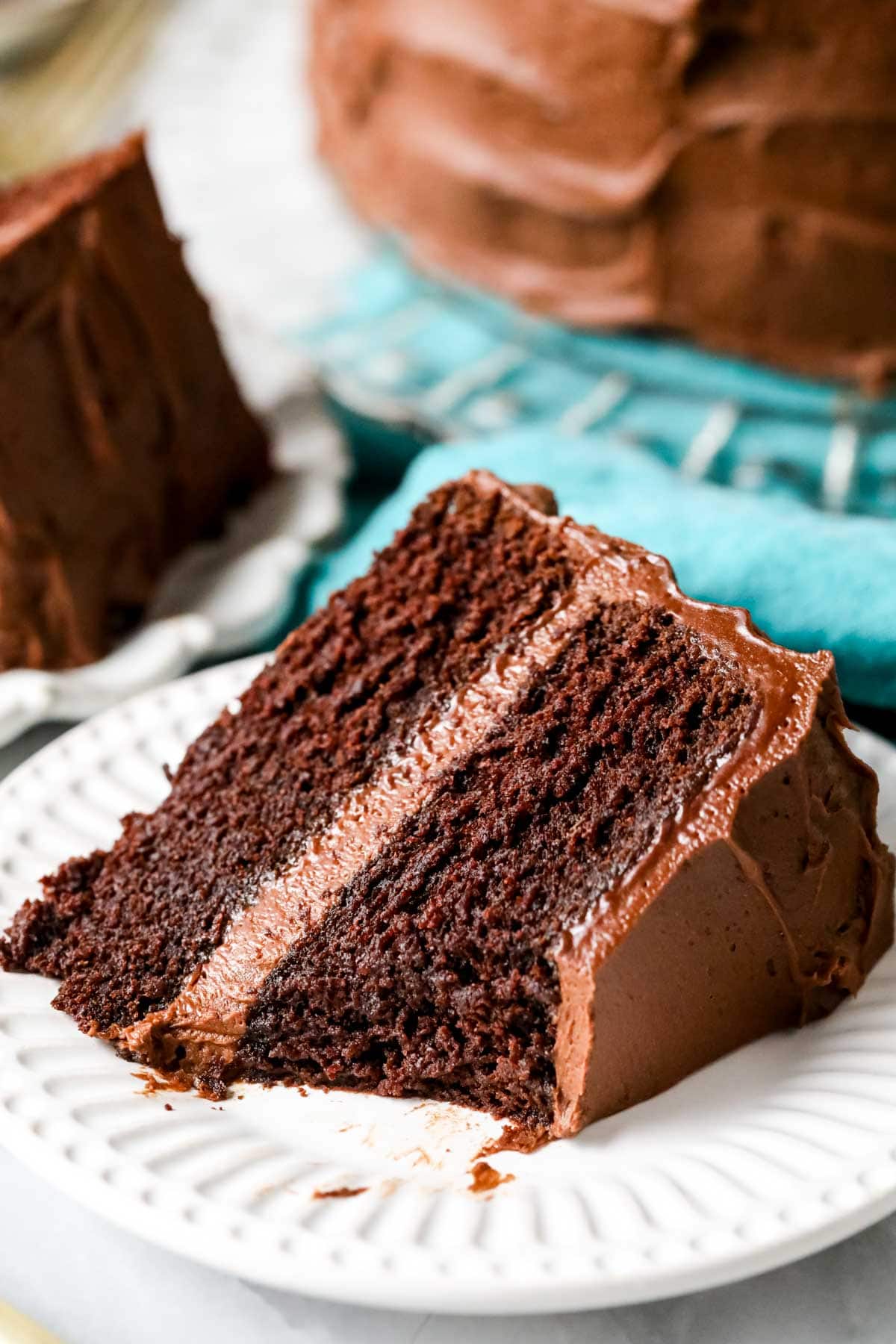 Slice of fudgy chocolate cake on a white plate with cake on crystal platter over blue cloth in background