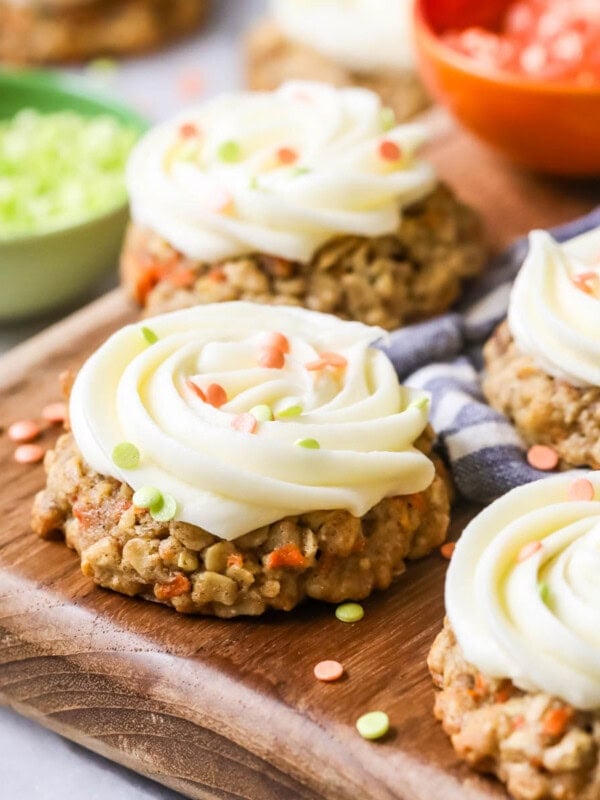 Carrot cake cookies toped with cream cheese frosting and sprinkles on a wood cutting board.