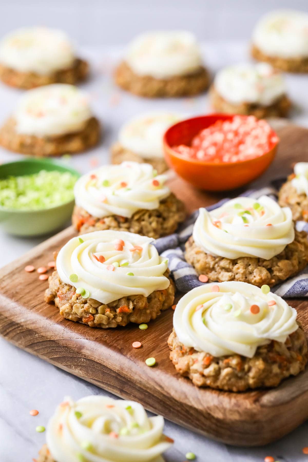 Carrot cake cookies toped with cream cheese frosting and sprinkles on a wood cutting board.