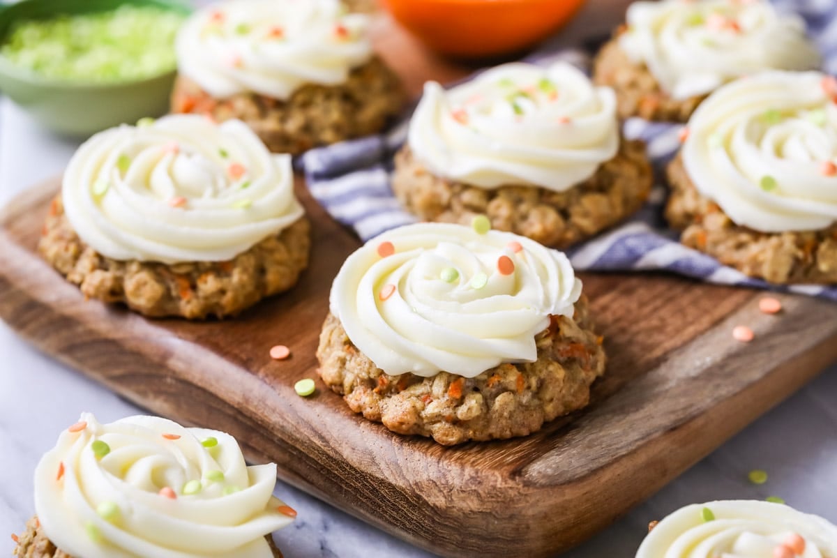 Carrot cake cookies toped with cream cheese frosting and sprinkles on a wood cutting board.
