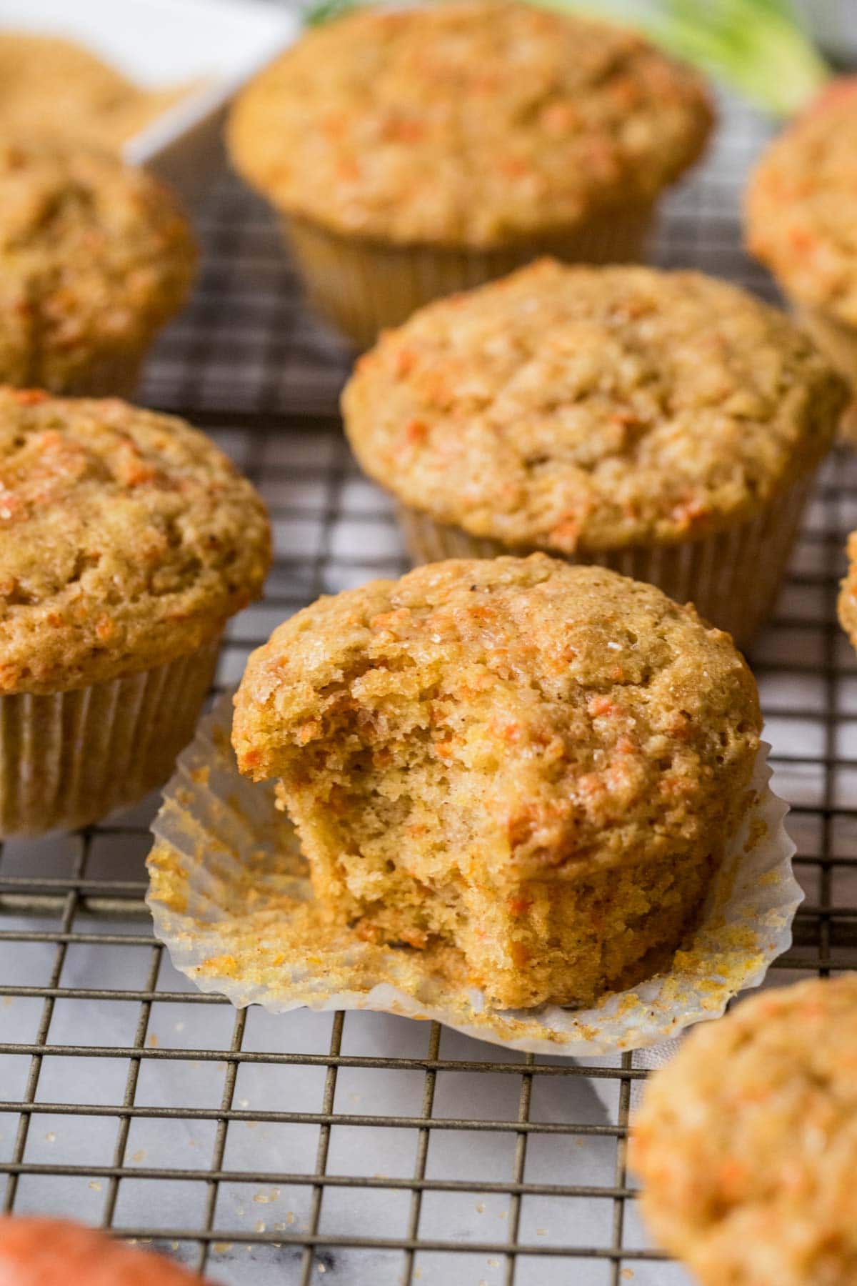 Carrot muffins on a cooling rack with the center muffin unwrapped and missing a bite.