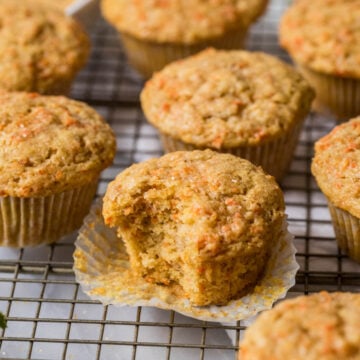 Carrot muffins on a cooling rack with the center muffin unwrapped and missing a bite.