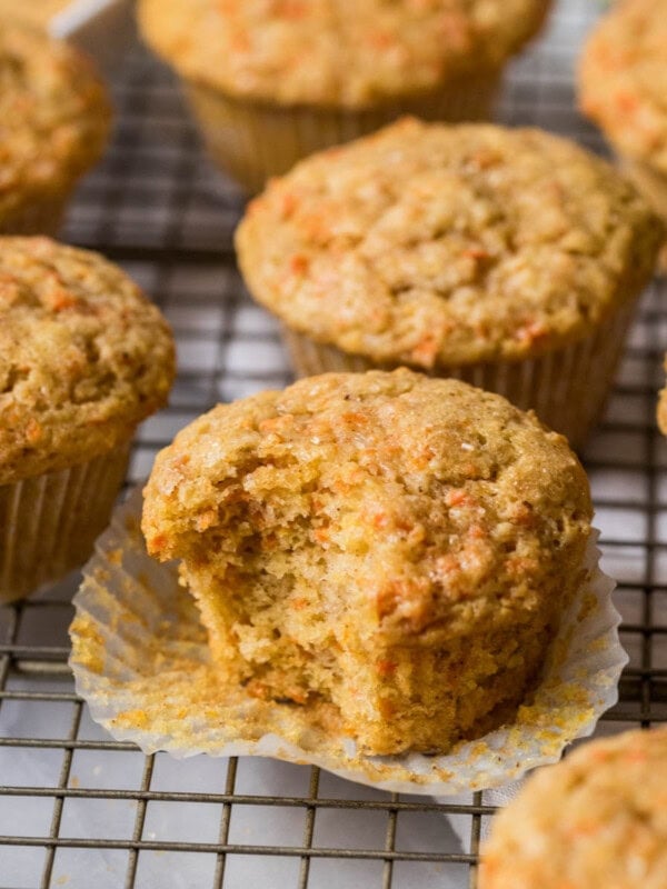 Carrot muffins on a cooling rack with the center muffin unwrapped and missing a bite.