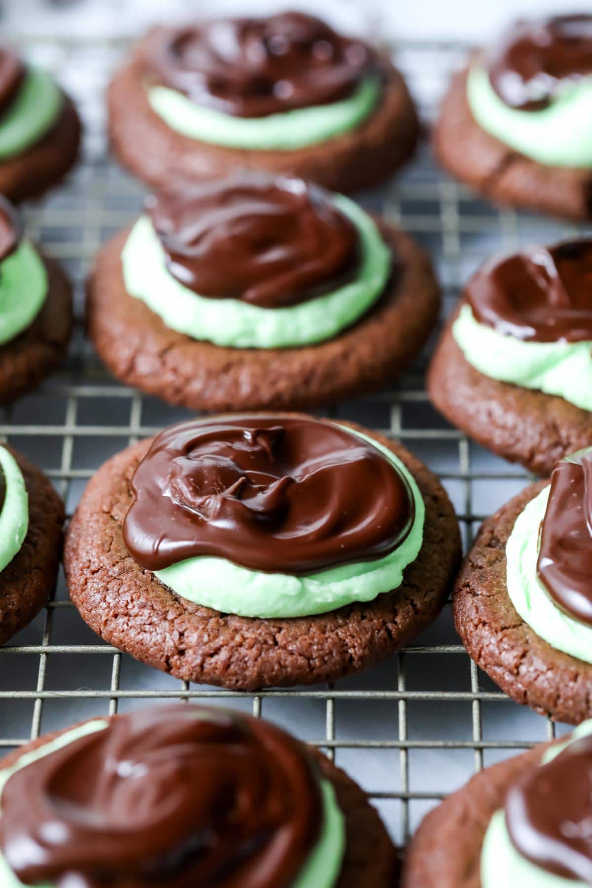 Mint chocolate cookies made with a chocolate cookie base, mint frosting, and chocolate ganache on a cooling rack.