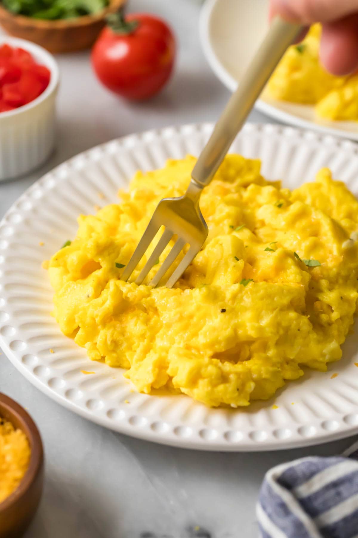 Fork diving into a pile of fluffy scrambled eggs on a white plate.