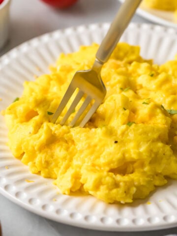 Fork diving into a pile of fluffy scrambled eggs on a white plate.