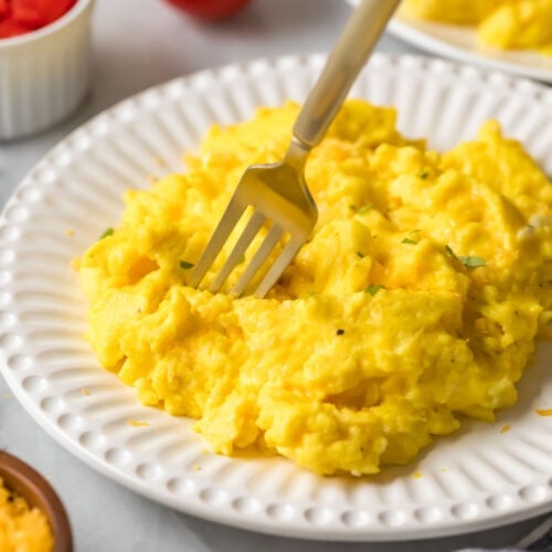 Fork diving into a pile of fluffy scrambled eggs on a white plate.