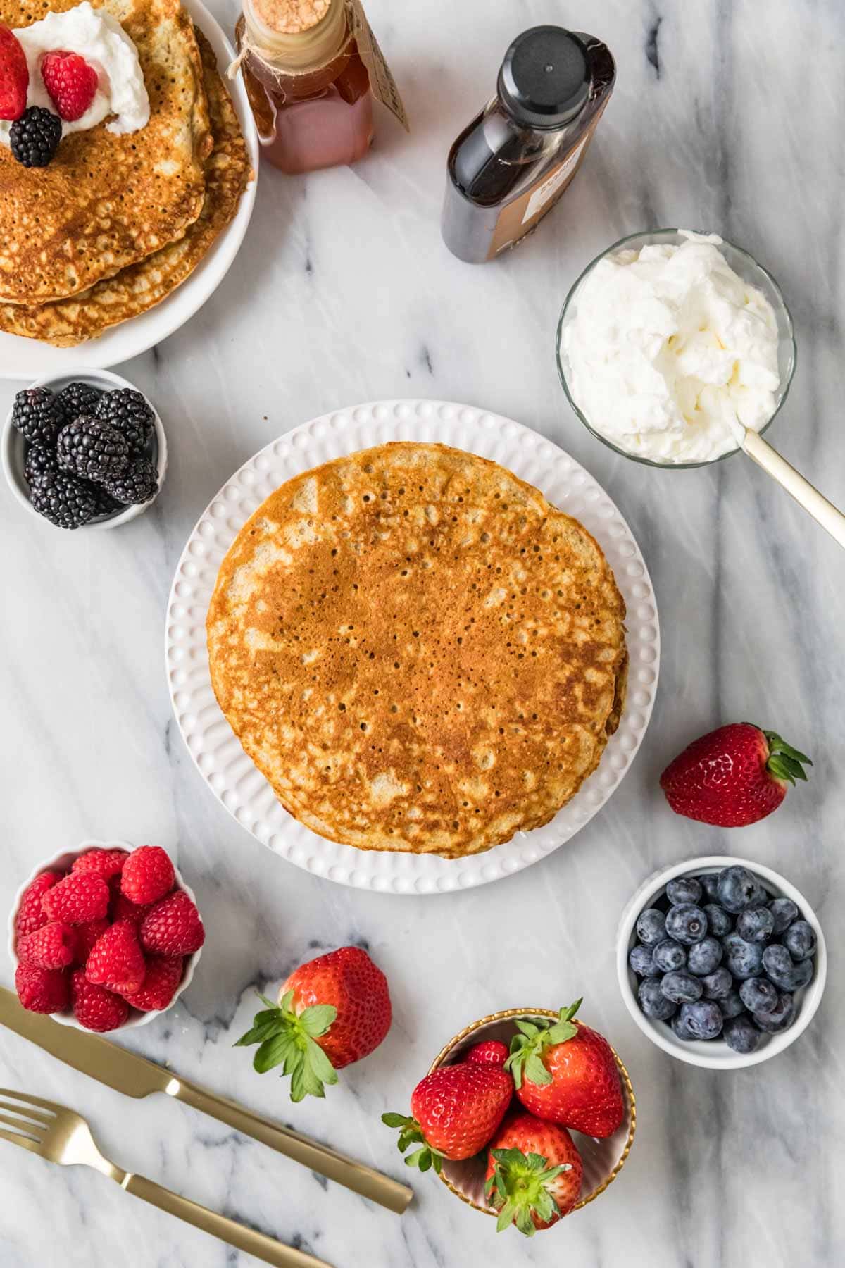 Overhead view of a whole wheat pancake on a plate surrounded by bowls of berries and whipped cream.