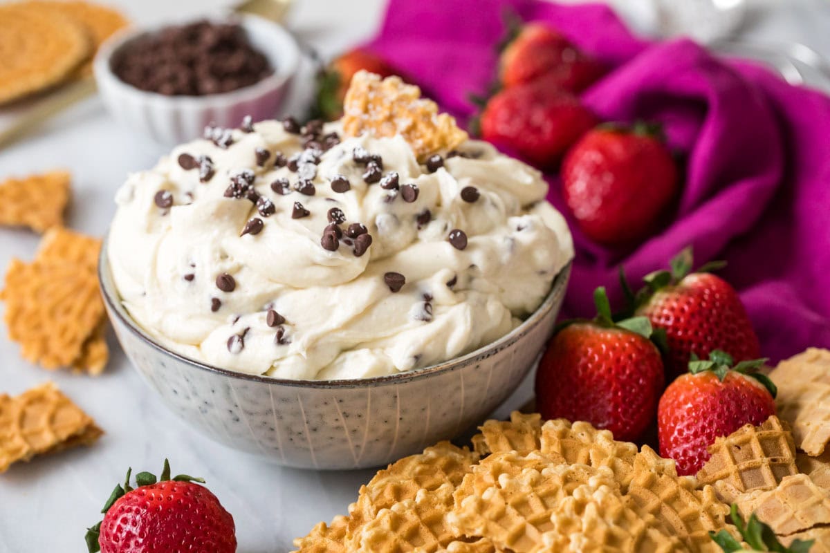 Bowl of dessert dip resembling a cannoli filling surrounded by strawberries and pizzelle cookies.