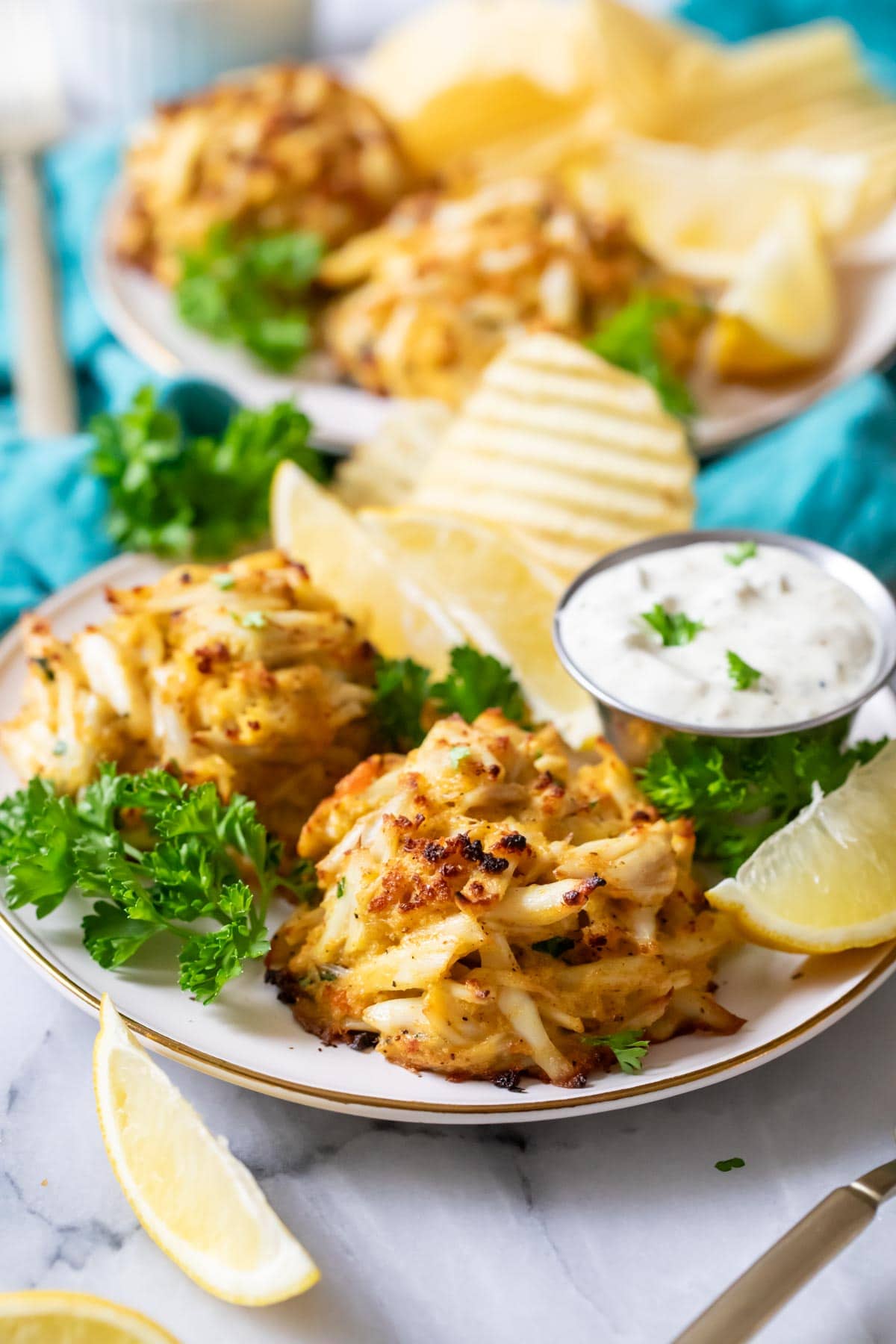 Plates with broiled crab cakes, tartar sauce, and ripped potato chips.