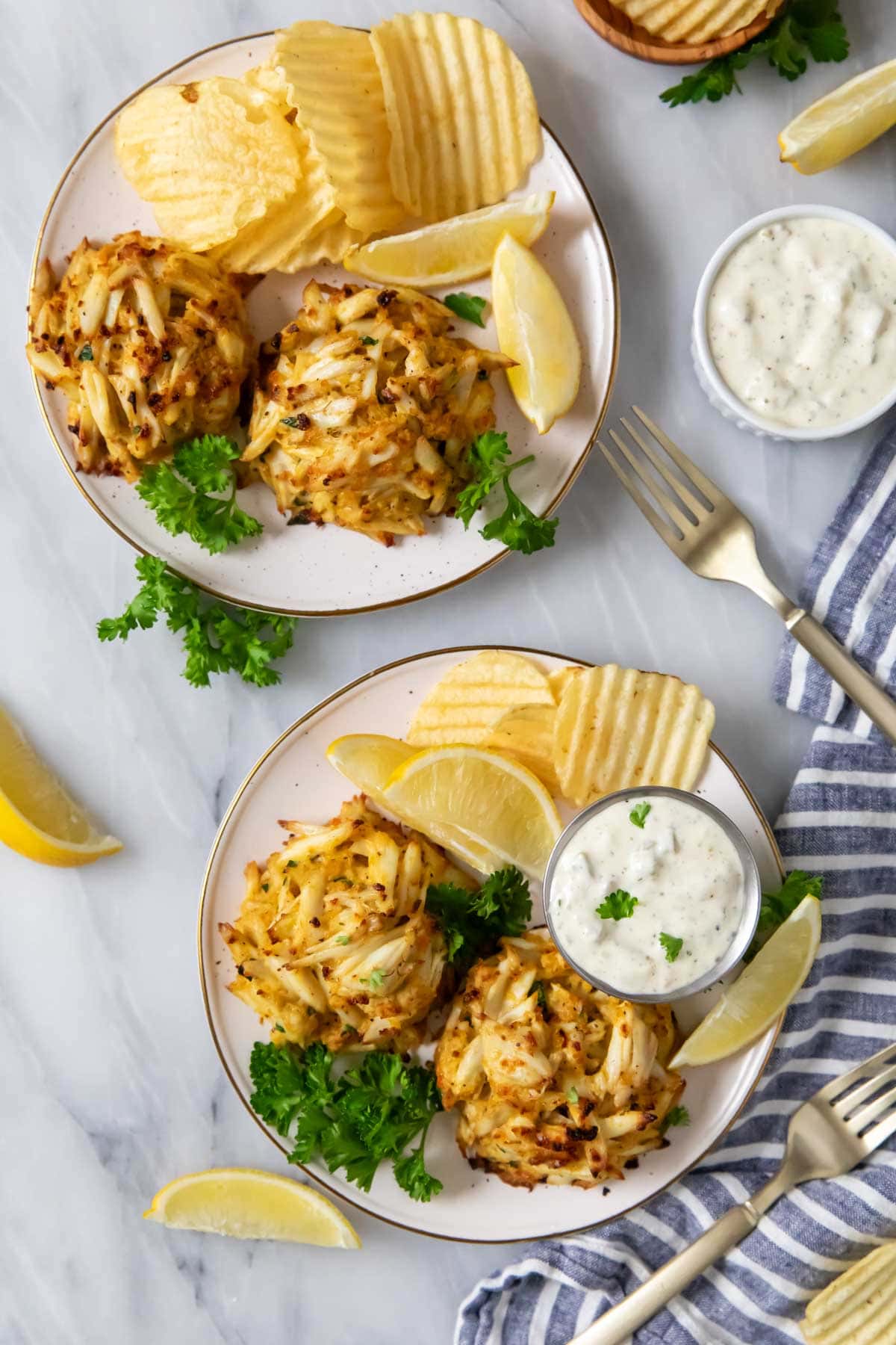 Overhead view of plates with broiled crab cakes, tartar sauce, and ripped potato chips.