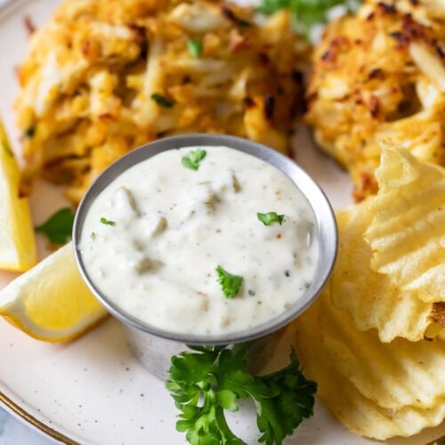 Dish of tartar sauce beside crab cakes and potato chips.