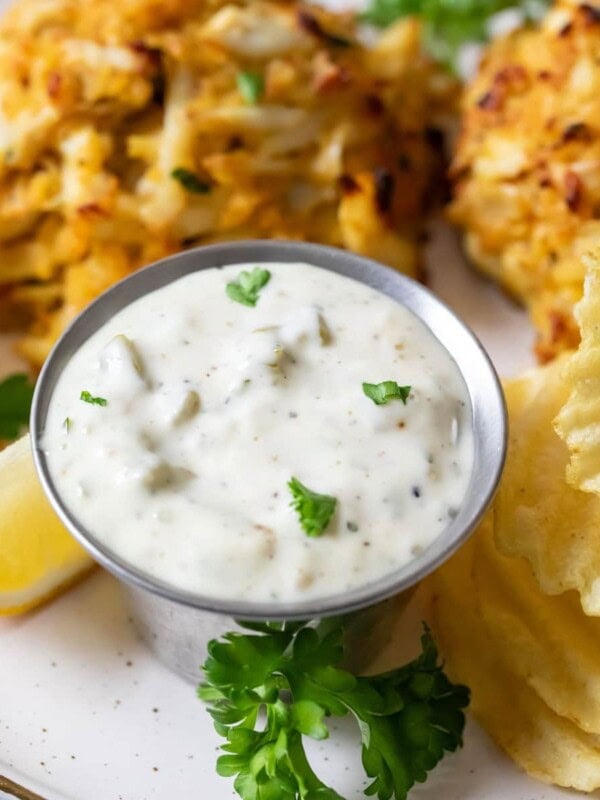 Dish of tartar sauce beside crab cakes and potato chips.