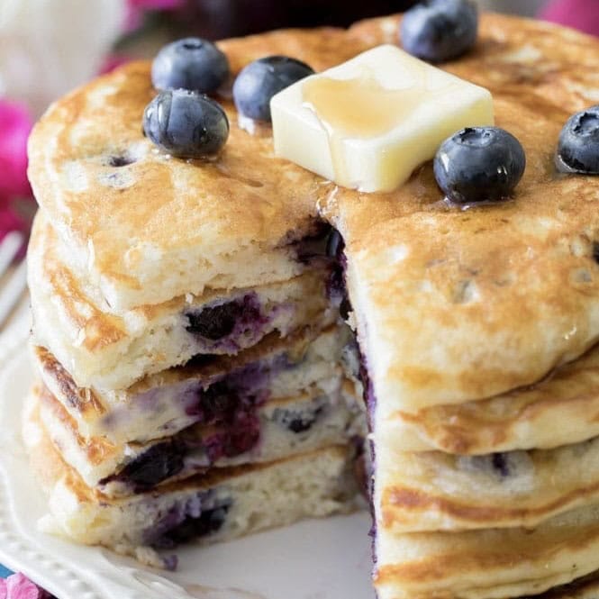 Stack of blueberry pancakes topped with blueberries, syrup, and a pat of butter.