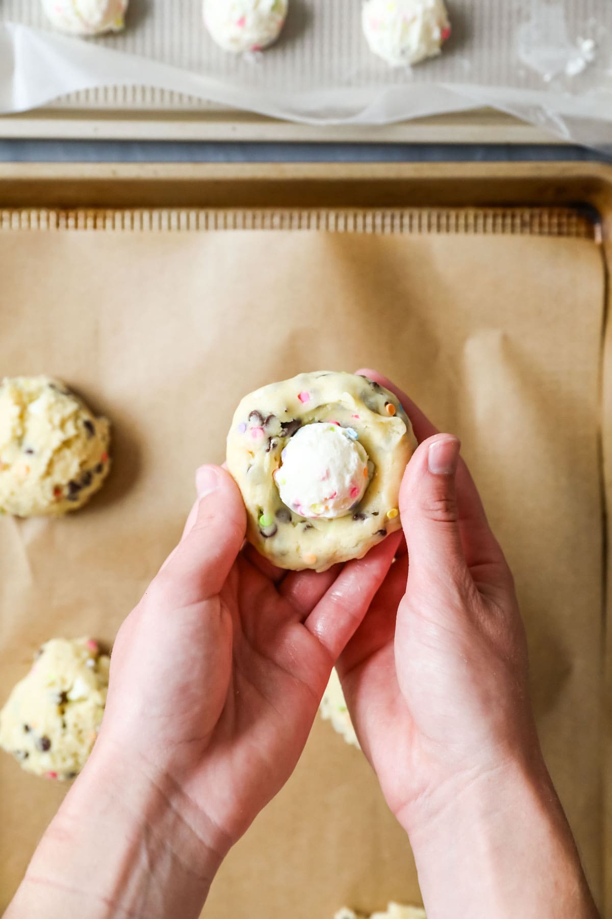 Hands showing cookie dough being folded around a buttercream ball.