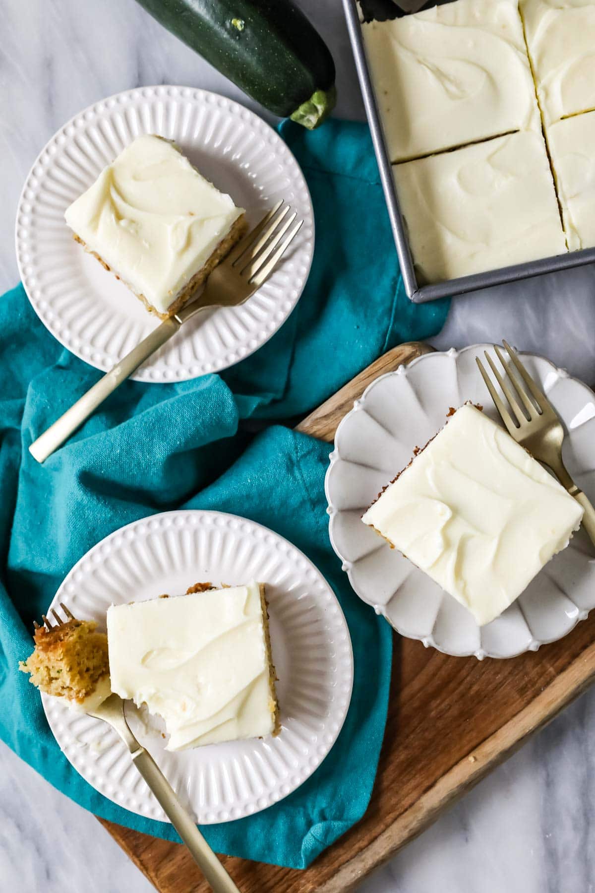 Overhead view of square slices of zucchini cake topped with cream cheese frosting on white plates.