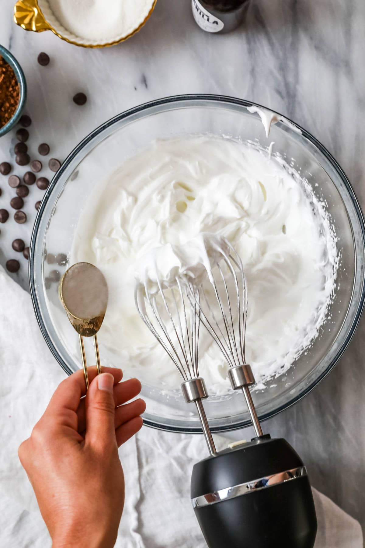 Overhead view of a bowl of sugar being added to egg whites whipped to soft peaks.