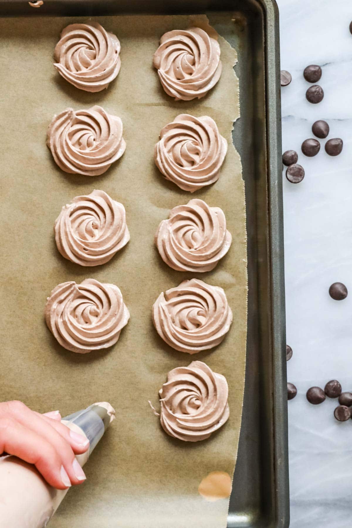 Overhead view of swirls of chocolate meringue being piped onto a parchment lined baking sheet.