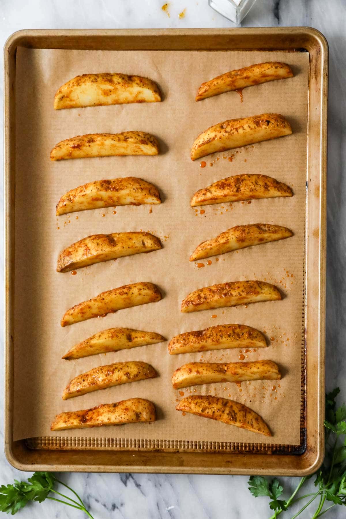 Overhead view of two neat rows of seasoned potato spears on a baking sheet.