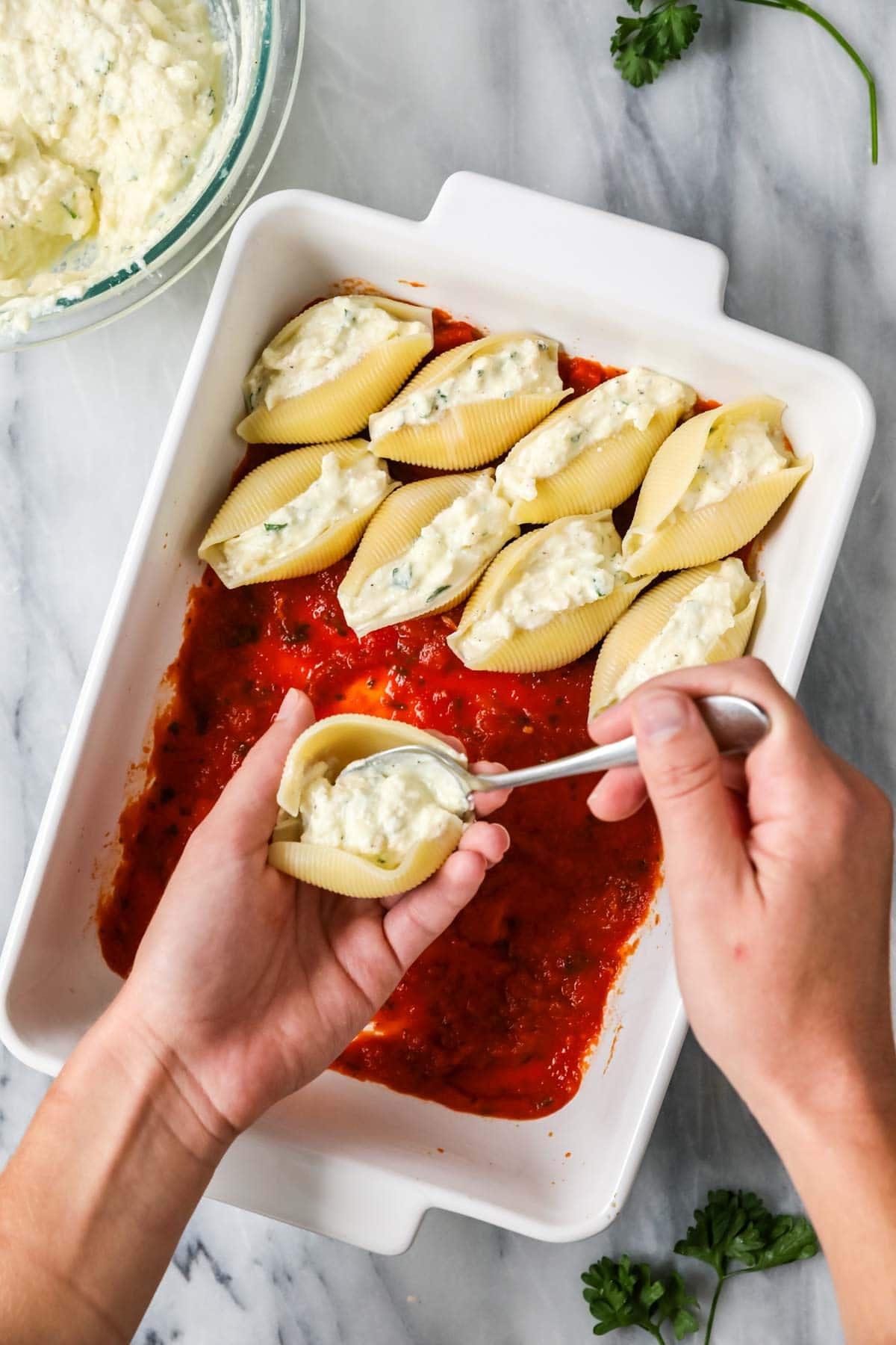 Overhead view of jumbo pasta shells being filled with a cheese filling and placed in a casserole dish with tomato sauce.