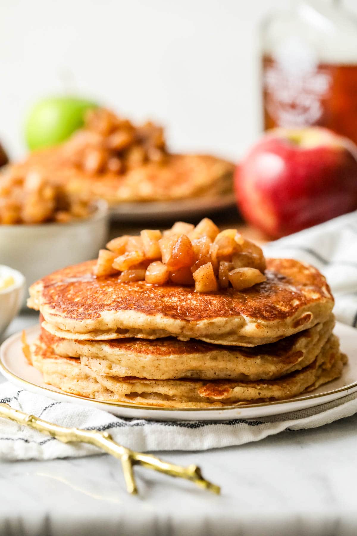 stack of apple pancakes on a white plate, syrup, more pancakes and apples in background