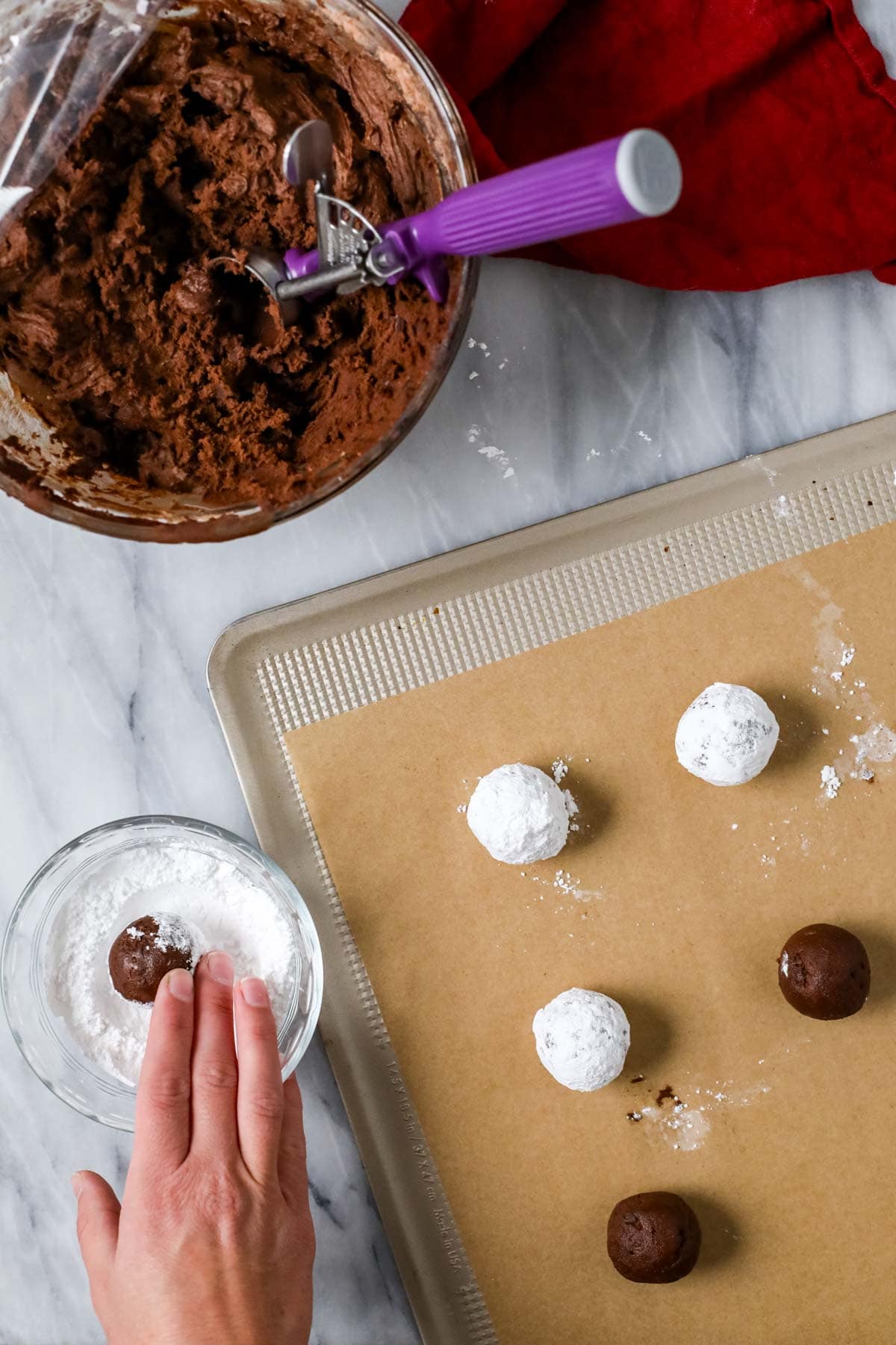 Hands rolling chocolate cookie dough balls through powdered sugar before baking.