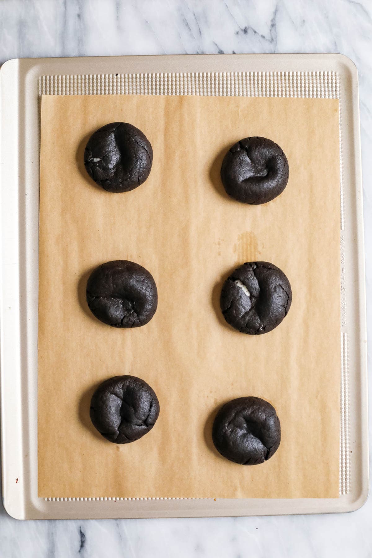 Overhead view of six cookies made with black cocoa on a parchment lined baking sheet.