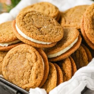 Pile of gingerbread sandwich cookies in a parchment lined metal tin.