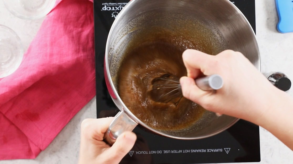 Whisk stirring a brown sugar meringue syrup in a mixer bowl over a burner.