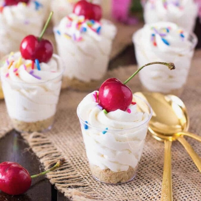Dessert shooter cups filled with cake batter cheesecake and topped with sprinkles and a cherry.