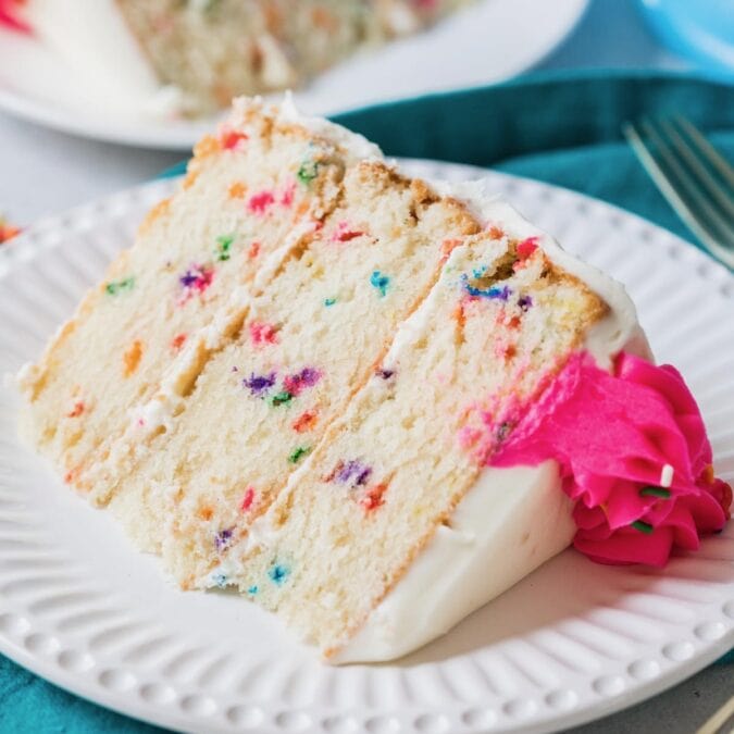 Slice of funfetti cake with lots of colorful sprinkles and a bright pink icing swirl on a white plate.