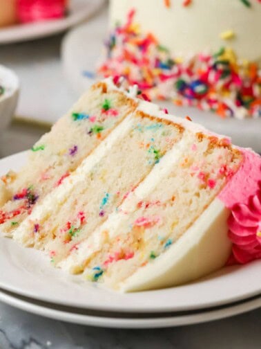 Slice of funfetti cake on a plate with the remaining cake in the background.