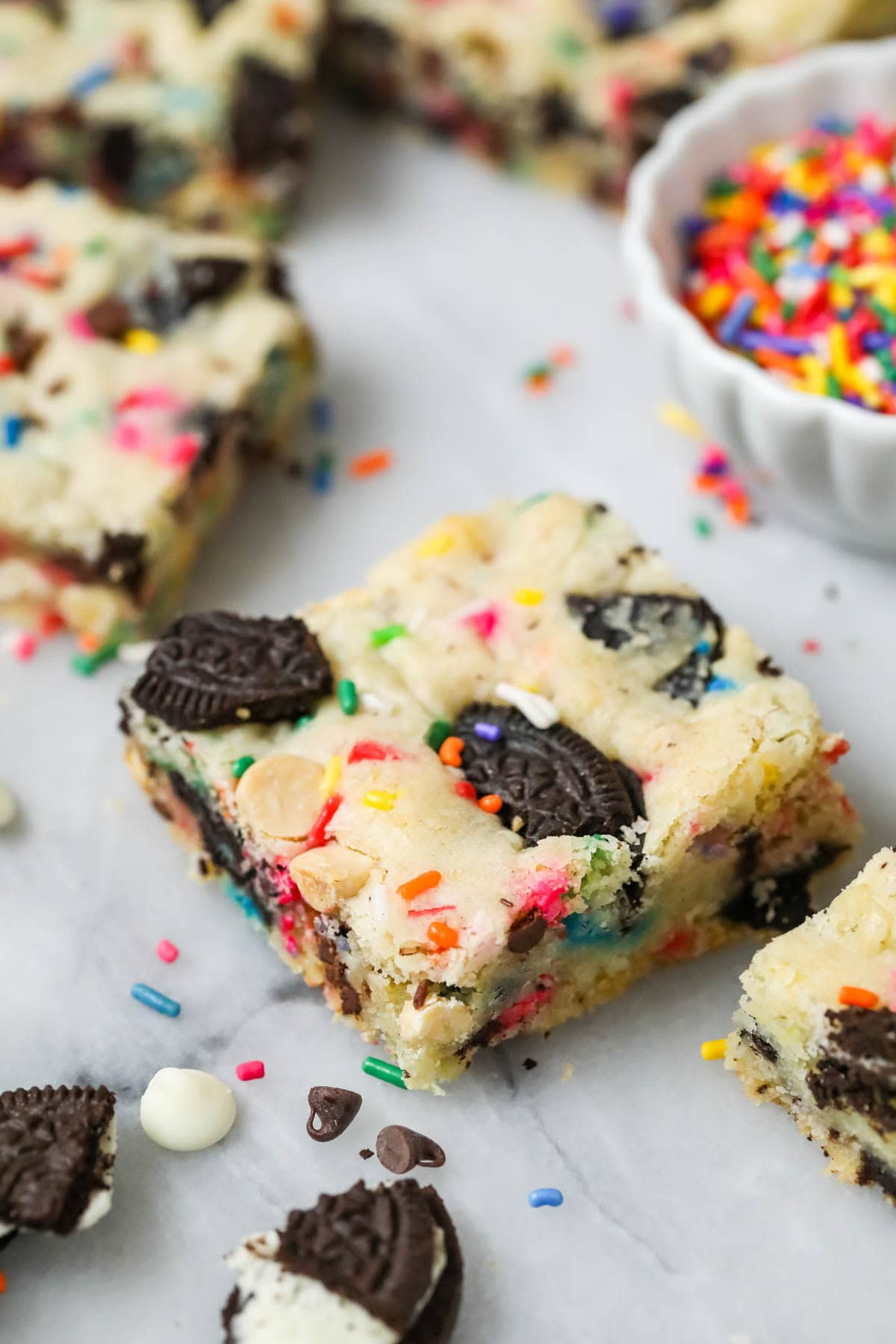 Funfetti Oreo bars on a marble surface with white chocolate chips, crushed Oreos, and sprinkles scattered all around.