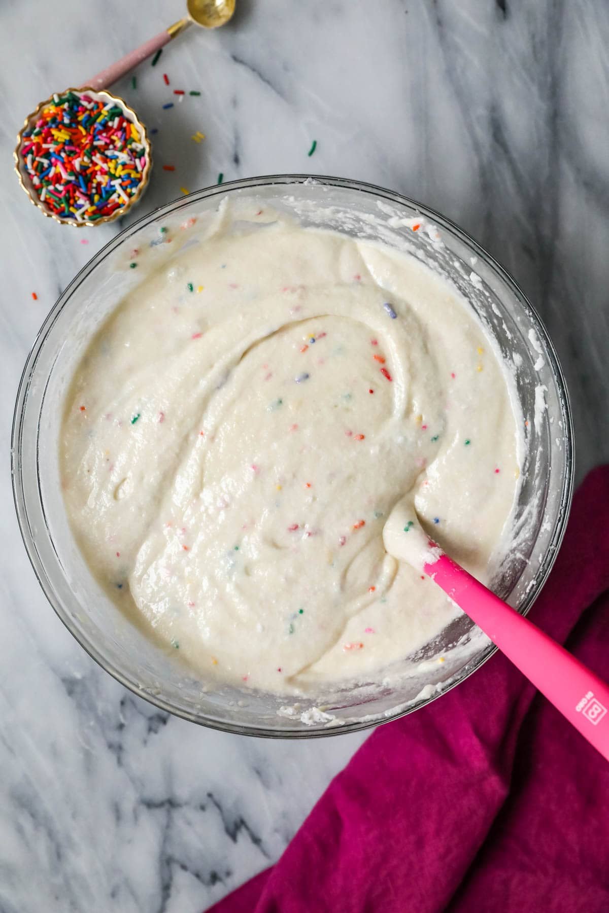 Overhead view of a bowl of sprinkle-flecked cake batter being stirred with a pink spatula.