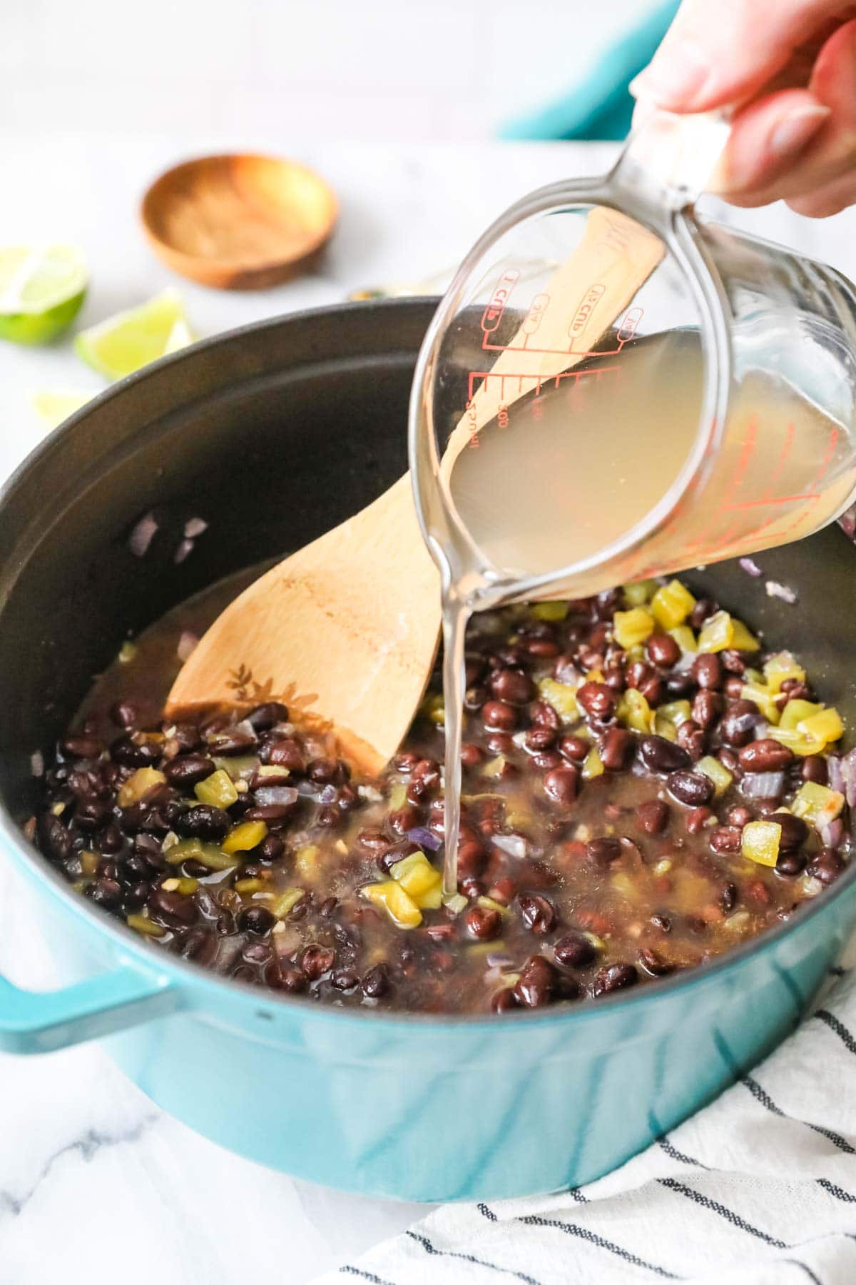 Pouring broth into a pot containing black beans, green chiles, and more.