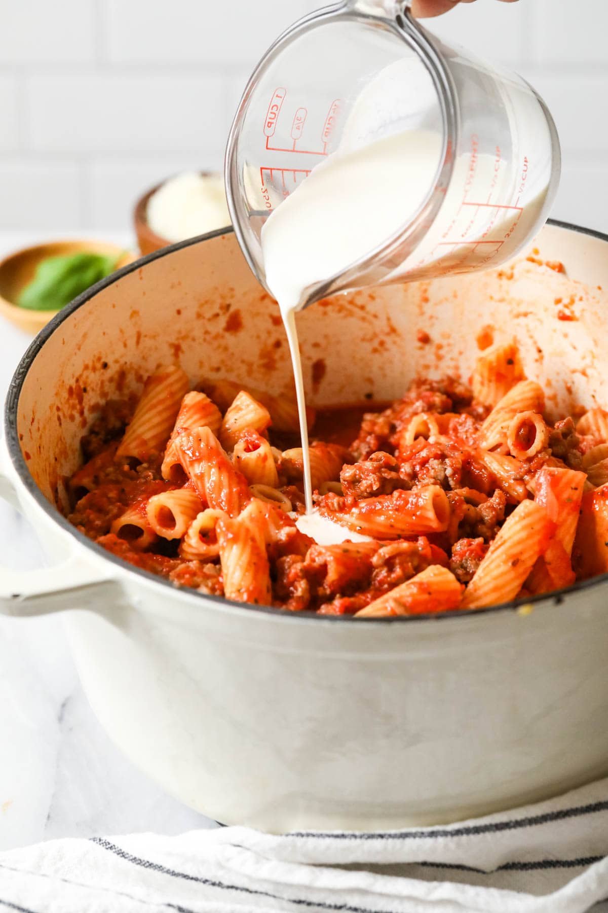 Cream being poured into a pot of rigatoni that's been tossed in a tomato and sausage sauce.