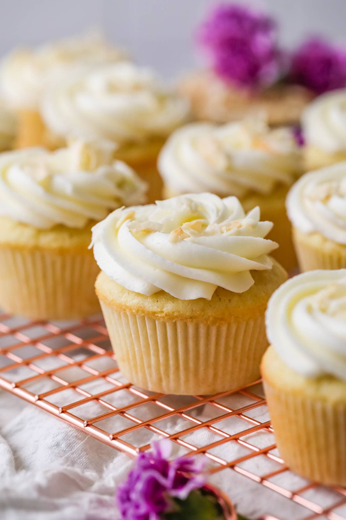 Rows of coconut cupcakes on a cooling rack with purple flowers in the background.