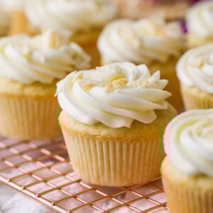 Rows of coconut cupcakes on a cooling rack with purple flowers in the background.
