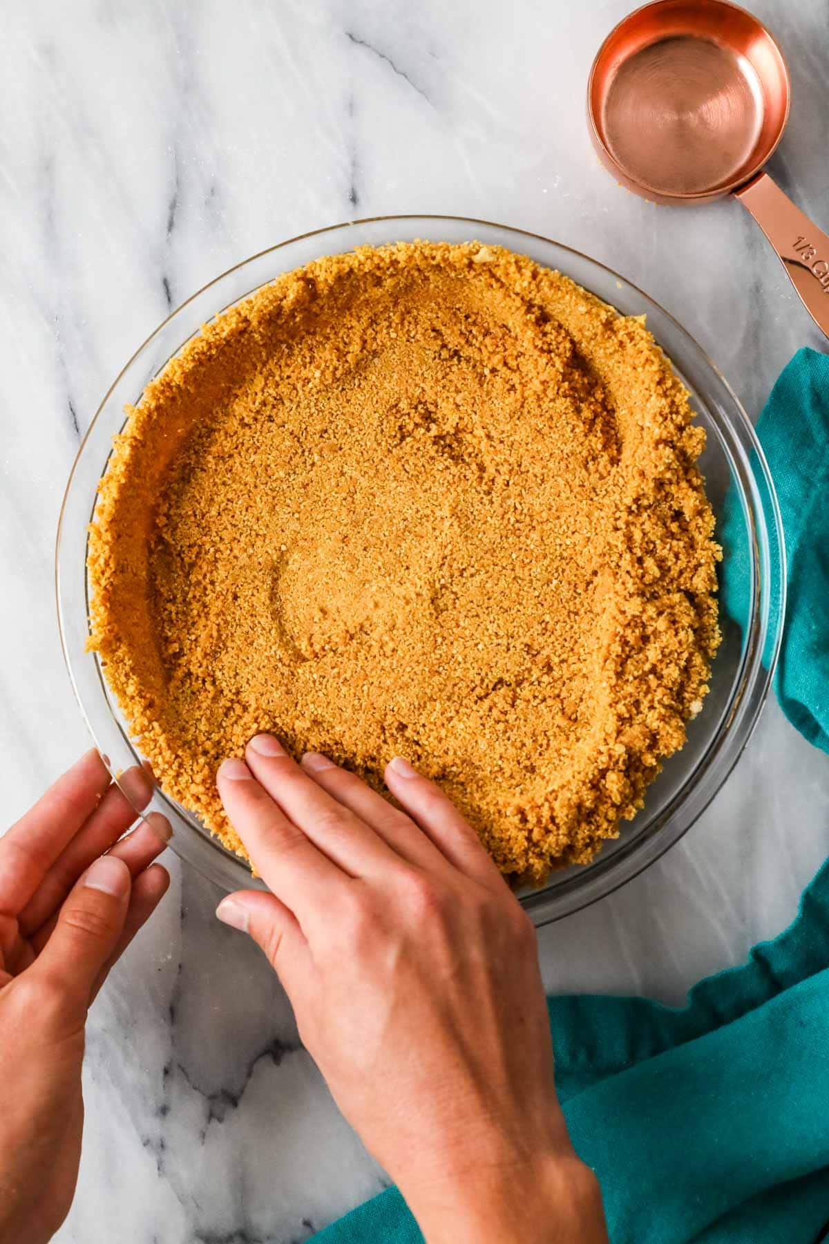Hands pressing graham cracker crumbs into a pie plate.