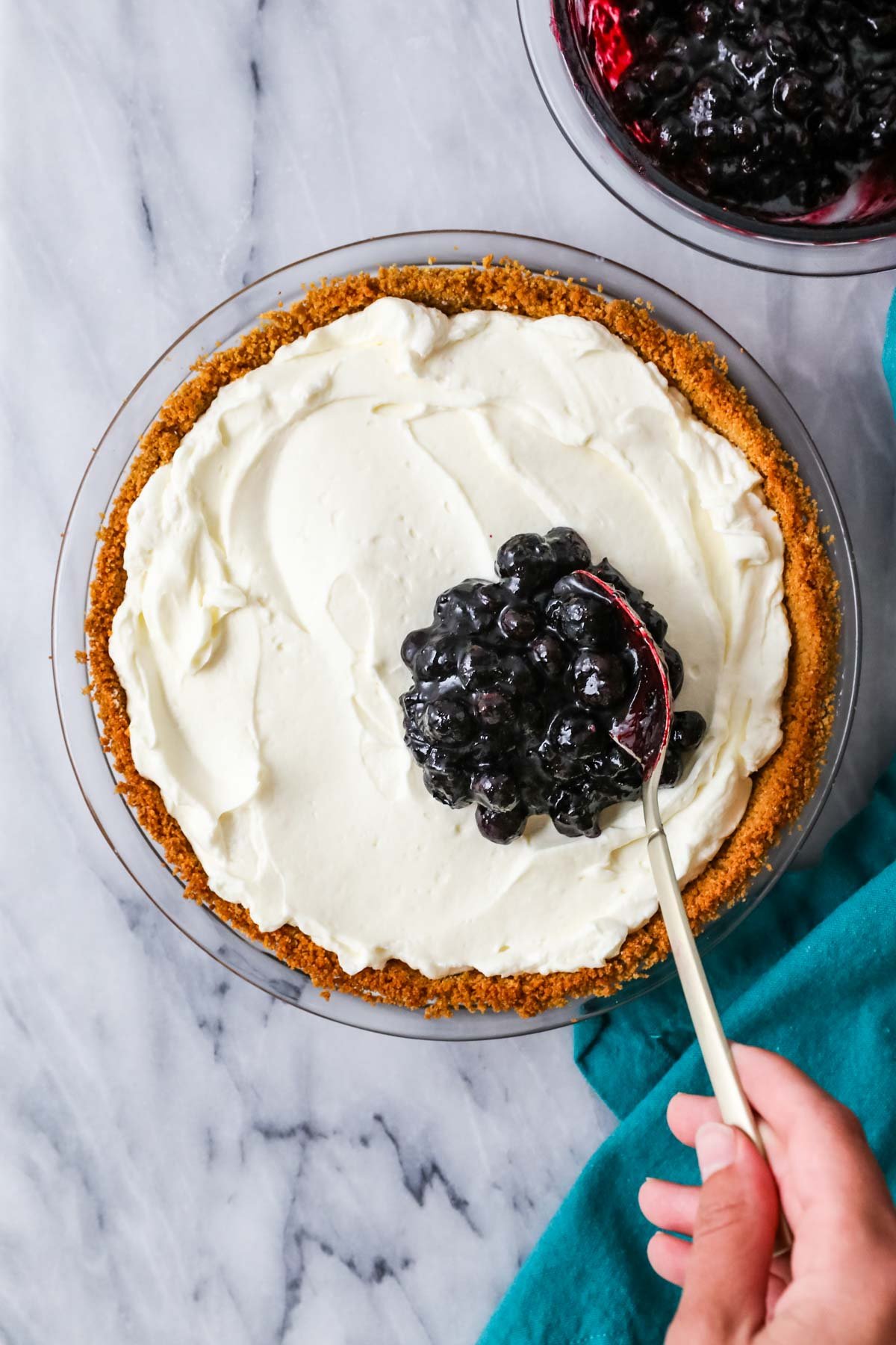 Blueberry topping being spooned over a cream cheese pie.