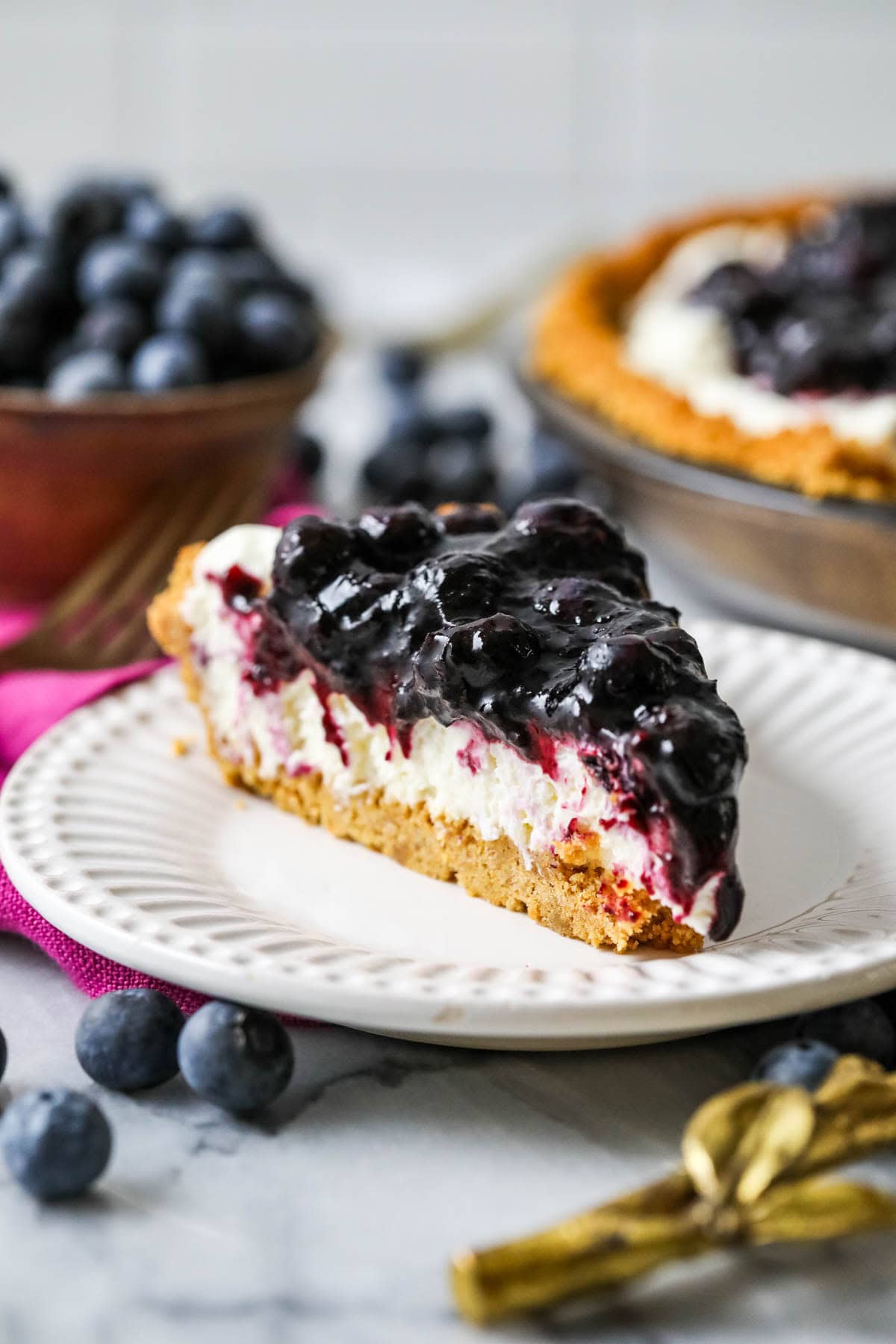 Slice of pie with a graham cracker crust, cream cheese filling, and blueberry topping.