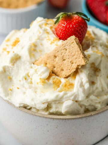 Bowl of cheesecake dip with graham cracker crumbs, a graham cracker, and a strawberry on top.