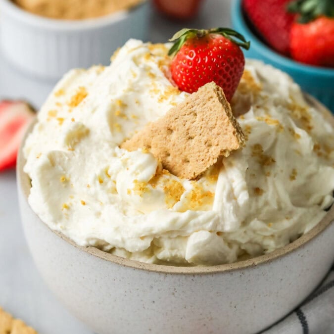 Bowl of cheesecake dip with graham cracker crumbs, a graham cracker, and a strawberry on top.