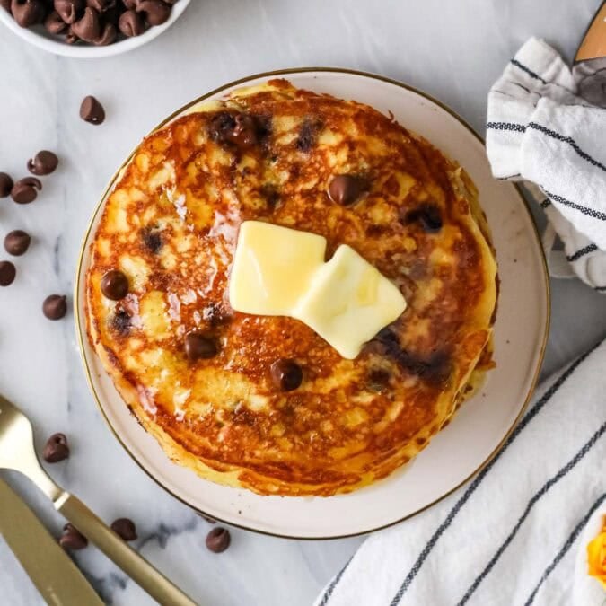 Overhead view of a stack of chocolate chip pancakes topped with butter and syrup.
