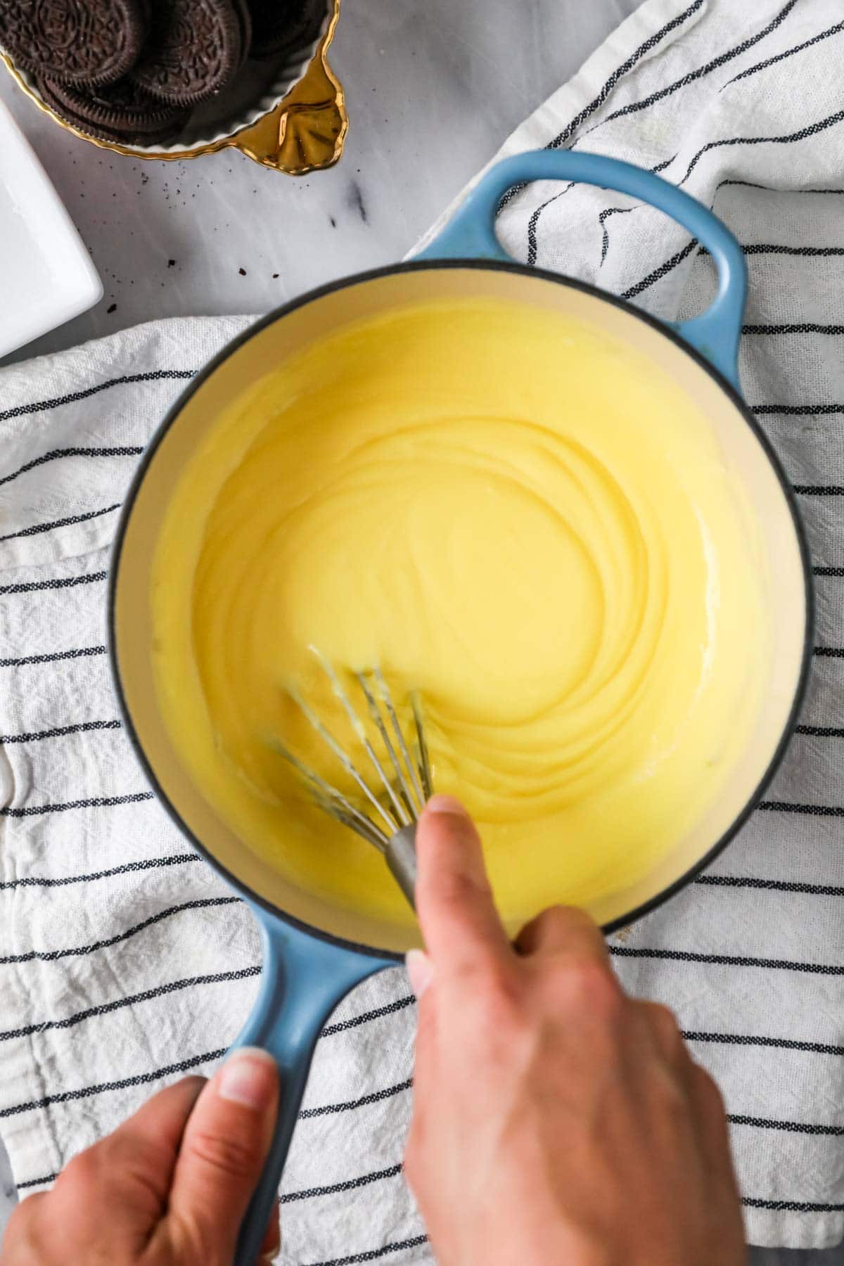 Homemade vanilla pudding in a blue saucepan, whisk is leaving trails in the pudding showing it's done.