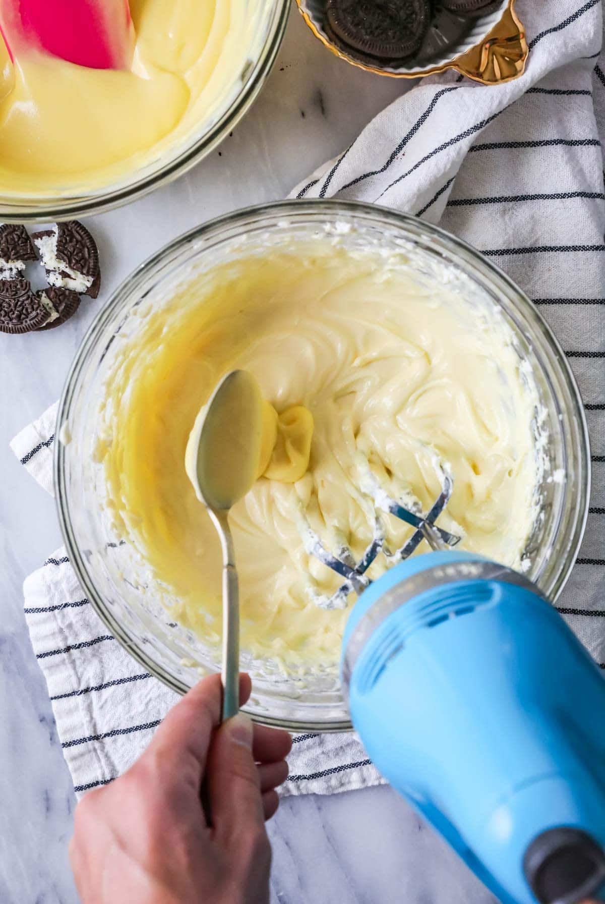Using a gold spoon to add pudding to the cream cheese mixture. and stirring with a blue mixer.