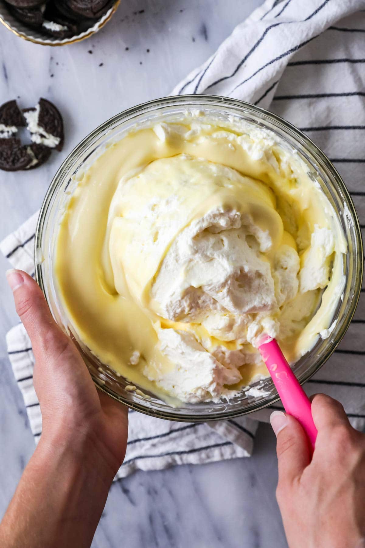 Folding whipped cream into pudding/cream cheese mixture in a glass bowl using a pink spatula.