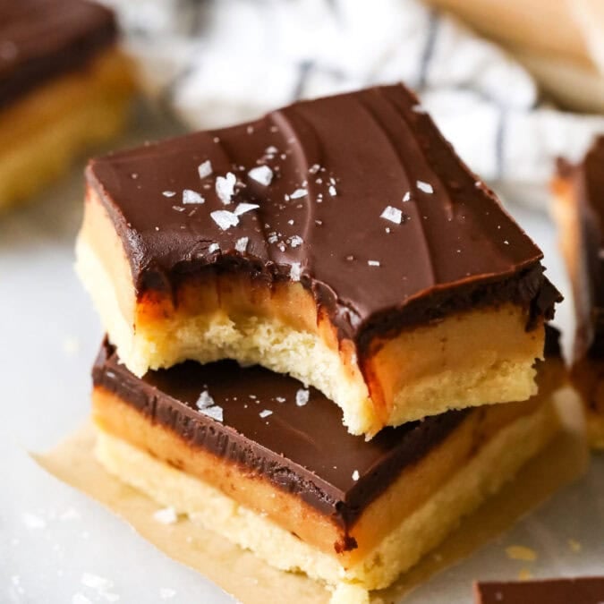Two square slices of millionaire's shortbread topped on top each other, a bite missing to show chewy caramel center. Topped with sea salt.