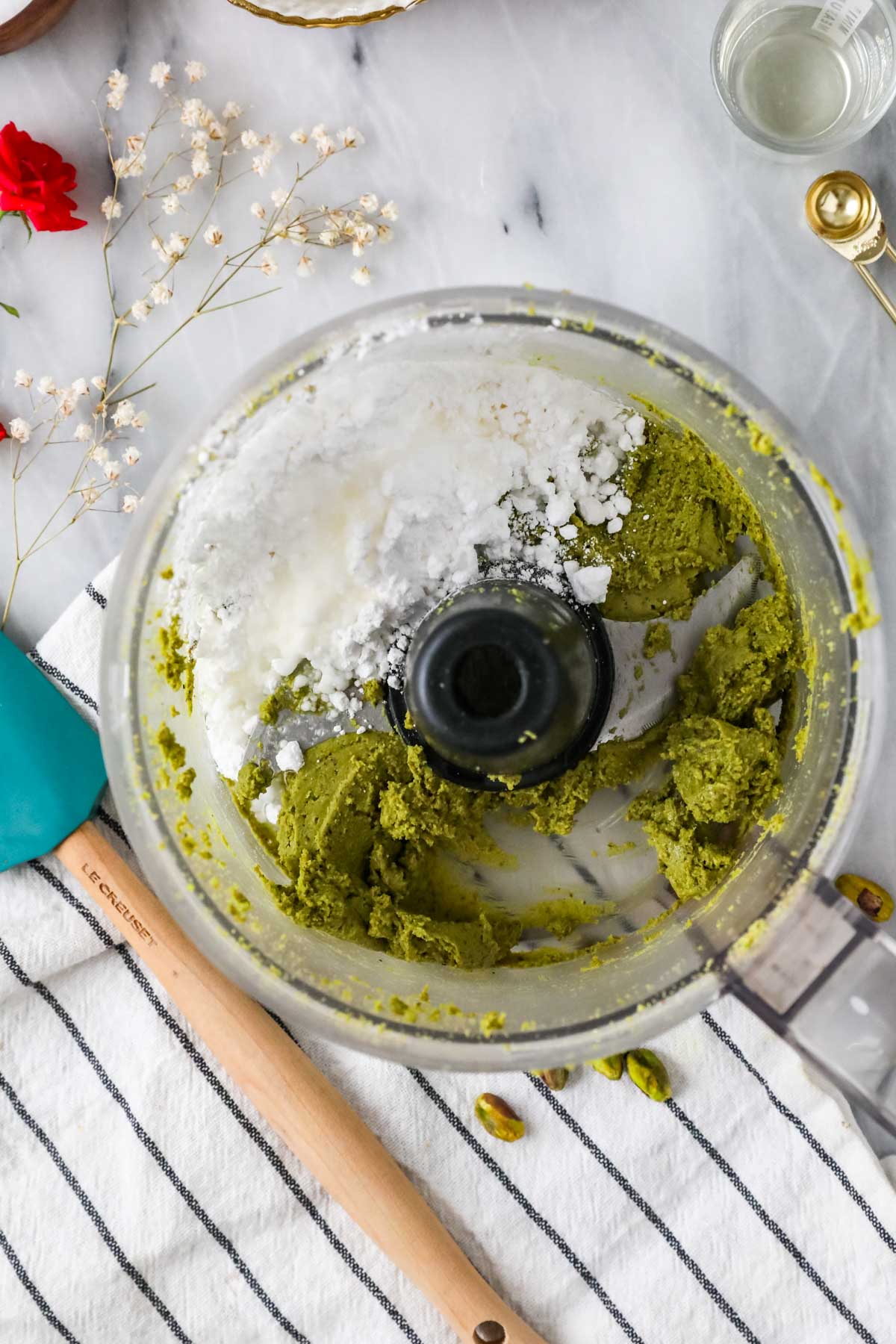 Overhead view of a food processor after adding powdered sugar, oil, and salt to ground pistachios.