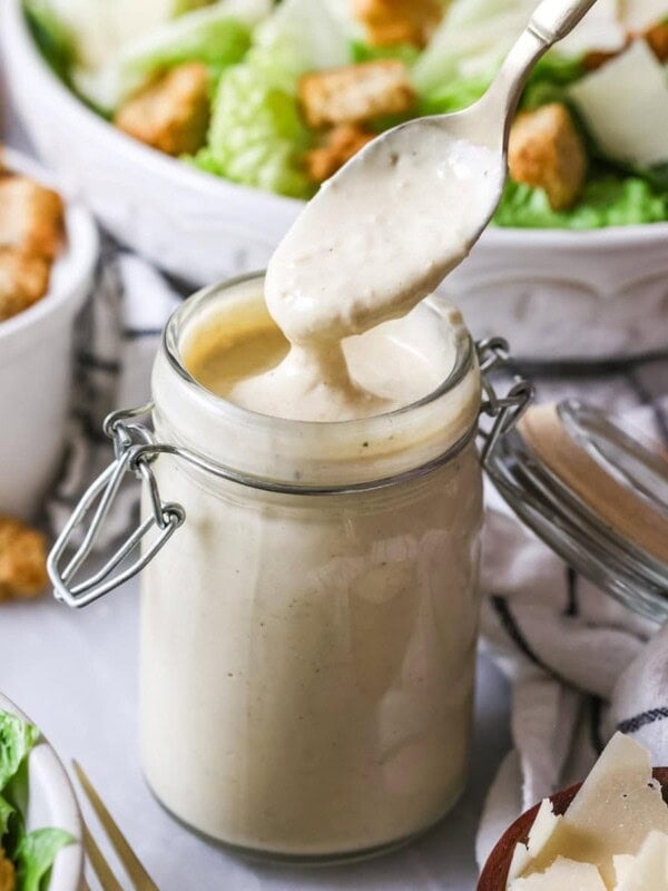 Spoon in a jar of homemade caesar dressing with salads in the background.